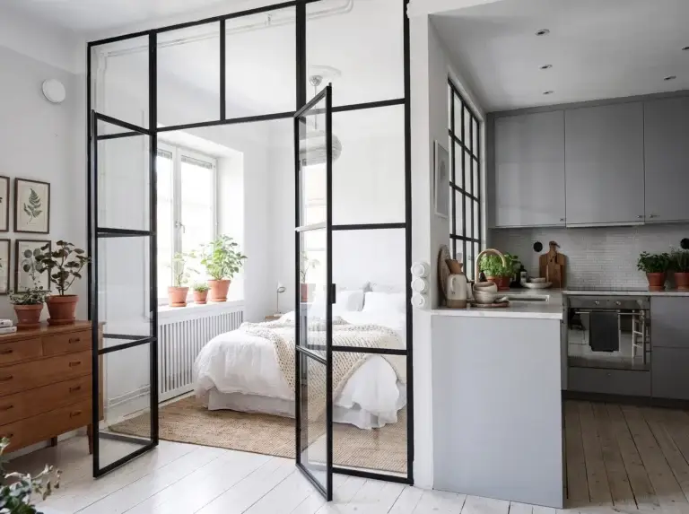 Black-framed glass wall with double doors separating a bedroom from a small kitchen