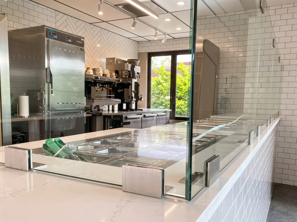 Frameless glass sneeze guard with stainless clamps over food pans at a cafe counter