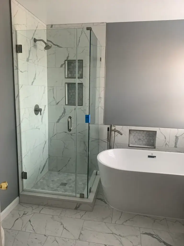 Frameless glass corner shower in marble-look tile beside a freestanding tub