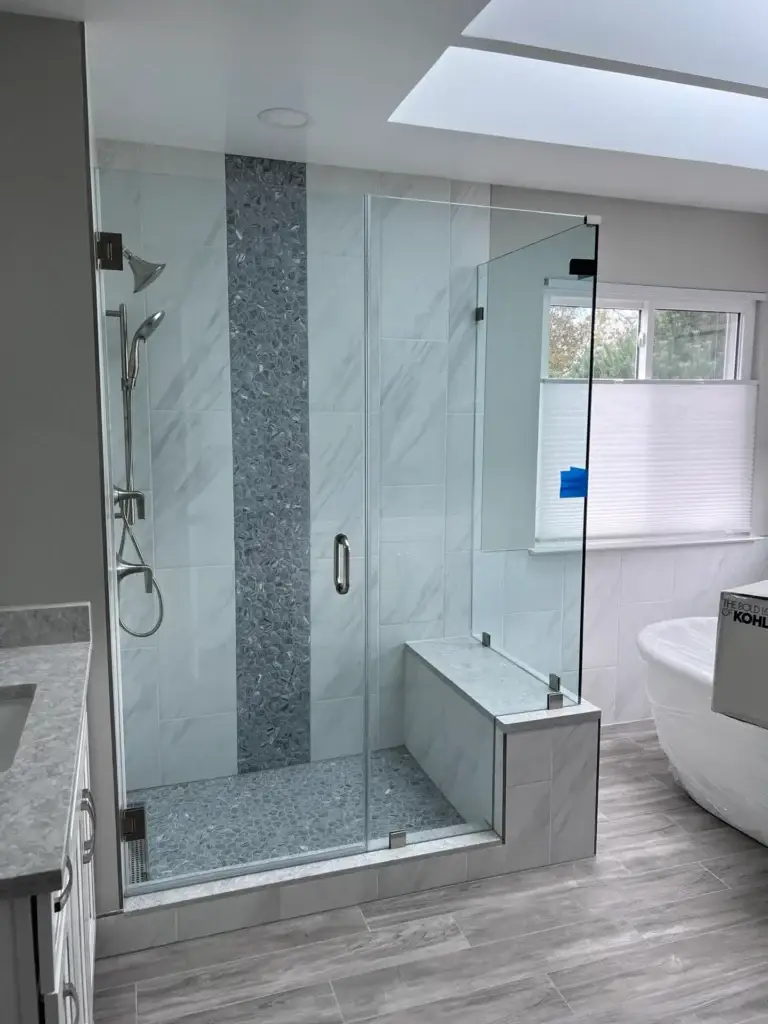 Frameless glass shower with a hinged door, bench and vertical mosaic accent under a skylight