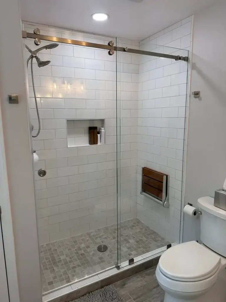 Frameless sliding shower door with a gold-tone rail in a white subway tile shower with a fold-down seat