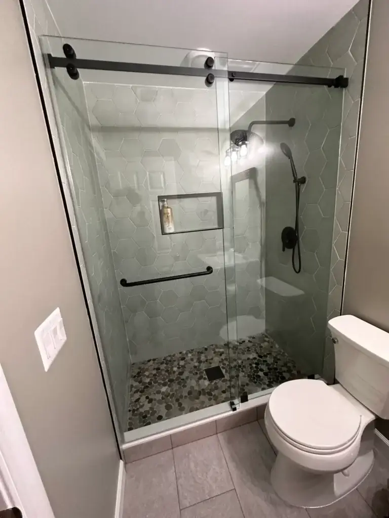 Frameless sliding shower door with matte black hardware in a grey tile shower with a pebble floor