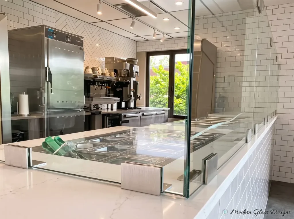 Frameless glass sneeze guard with stainless clamps over food pans at a cafe counter