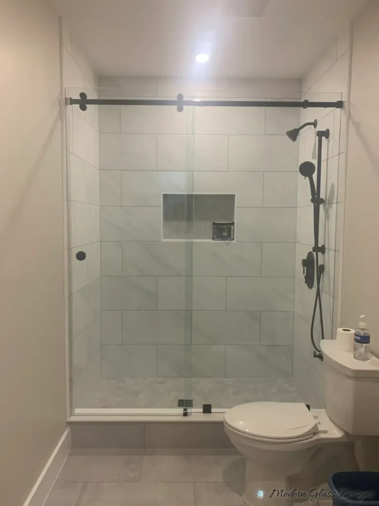 Frameless sliding shower door with a dark-finish rail in a white marble-look tile shower
