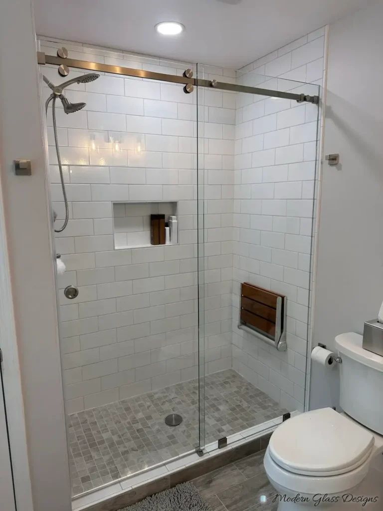 Frameless sliding shower door with a gold-tone rail in a white subway tile shower with a fold-down seat