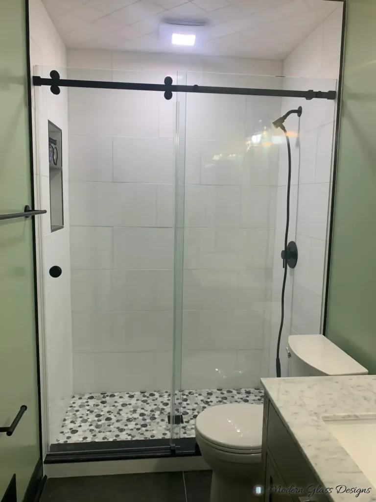 Frameless sliding shower door with a matte black rail in a white tile shower with a pebble floor