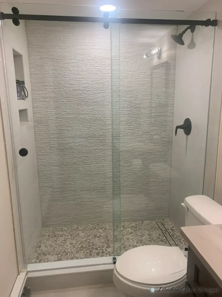 Frameless sliding shower door with matte black hardware, textured white tile and a pebble floor