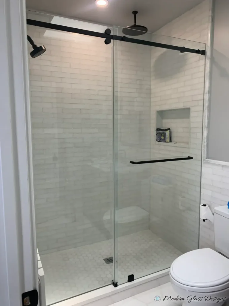 Frameless sliding shower door with a matte black rail and towel bar in a white subway tile shower
