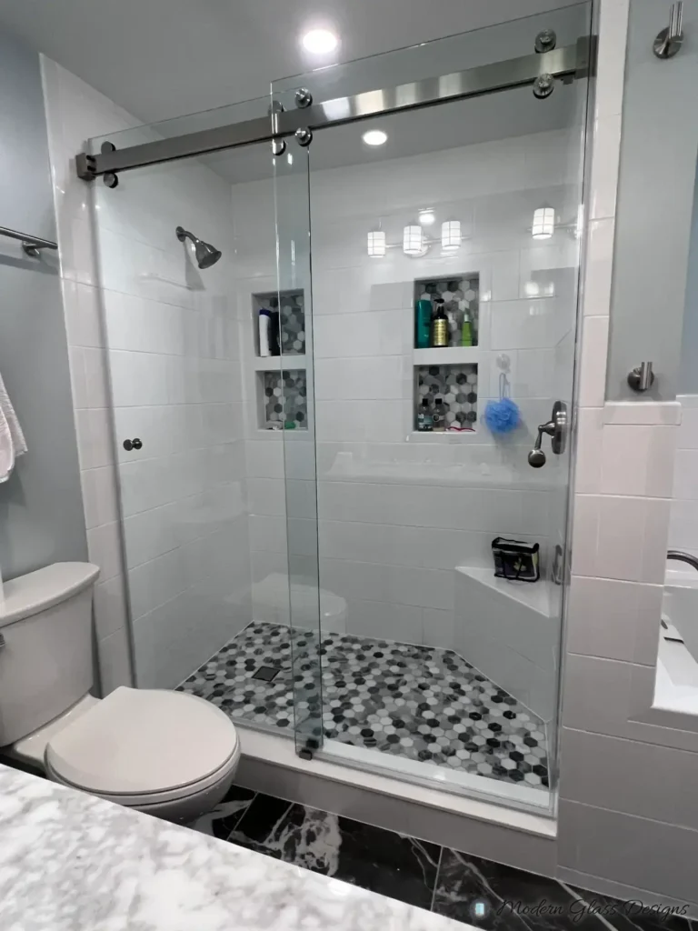 Frameless sliding shower door in a white tile shower with mosaic niches and a hex floor