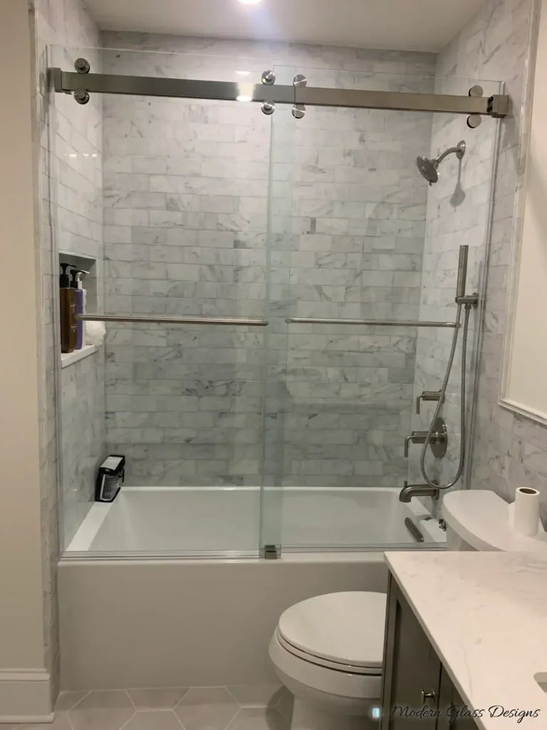 Frameless sliding glass doors with towel bars over a bathtub with marble subway tile