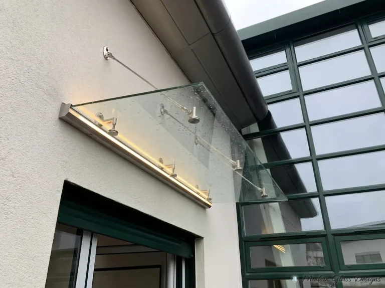 Glass canopy with a lit stainless wall rail and support rods above a side door